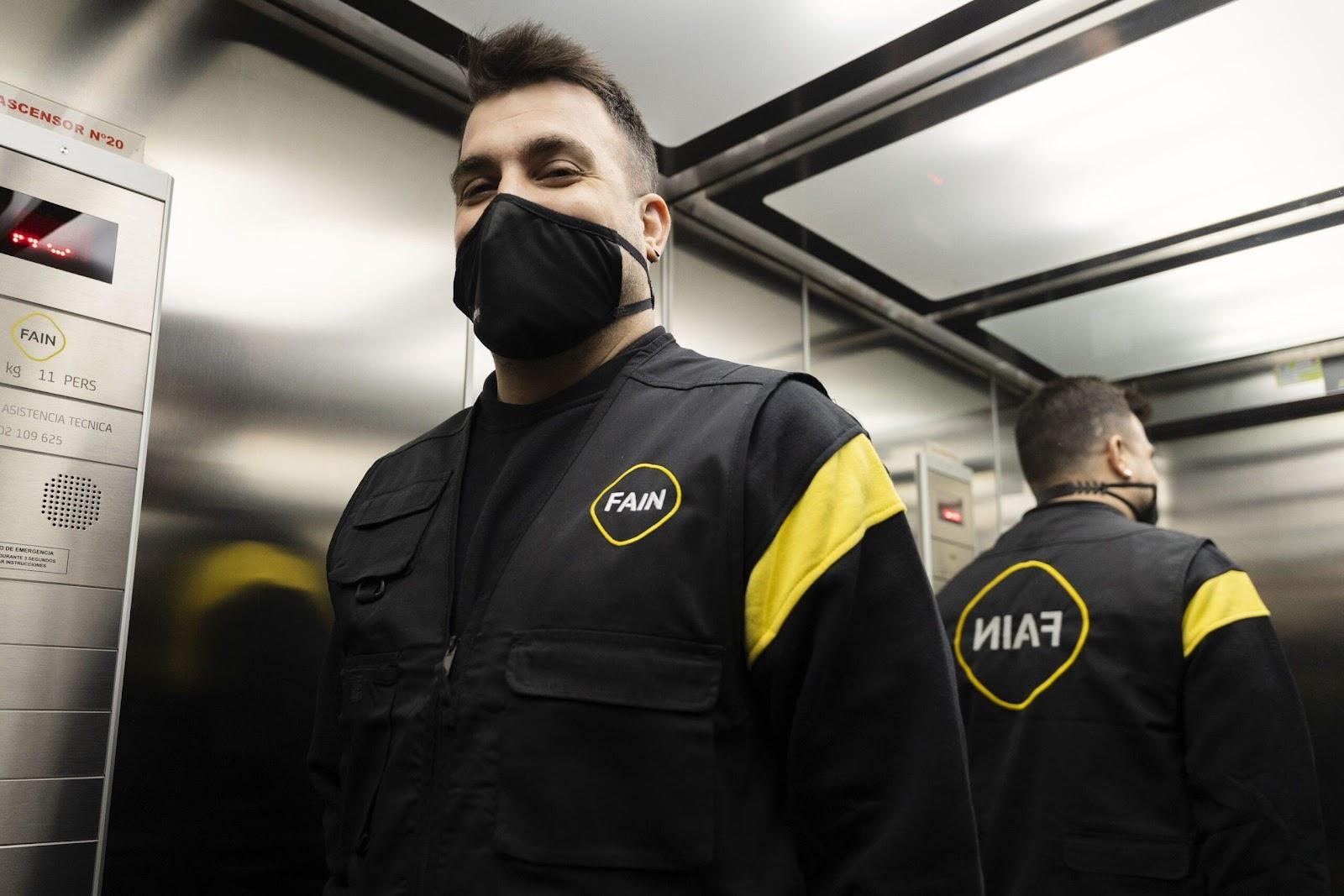 Technicien portant un uniforme noir et jaune FAIN et un masque, à l'intérieur d'un ascenseur, avec son reflet visible dans le miroir.